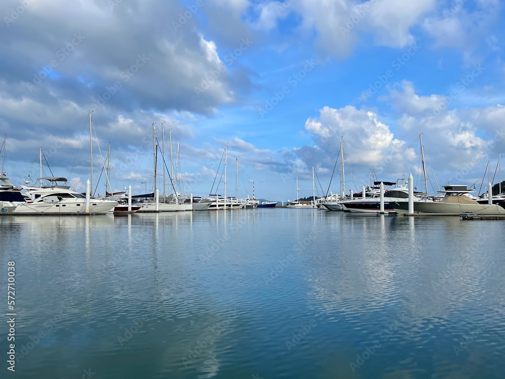 Beautiful view of luxury yachts in the harbor. Sea bay. Blue sky with clouds reflect on the surface of water. Boats at anchor. Sailboats with masts pointing to the sky. Yacht Club. Tropical paradise.