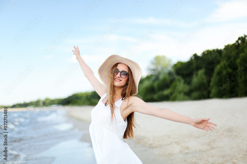 Happy smiling woman in free happiness bliss on ocean beach standing with a hat, sunglasses, and raising hands. Portrait of a multicultural female model in white summer dress enjoying nature Happy smiling woman in free happiness bliss on ocean beach standing with a hat, sunglasses, and raising hands. Portrait of a multicultural female model in white summer dress enjoying nature