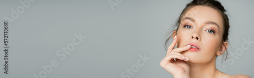 portrait of charming woman with natural makeup and hand near face looking at camera isolated on grey, banner.