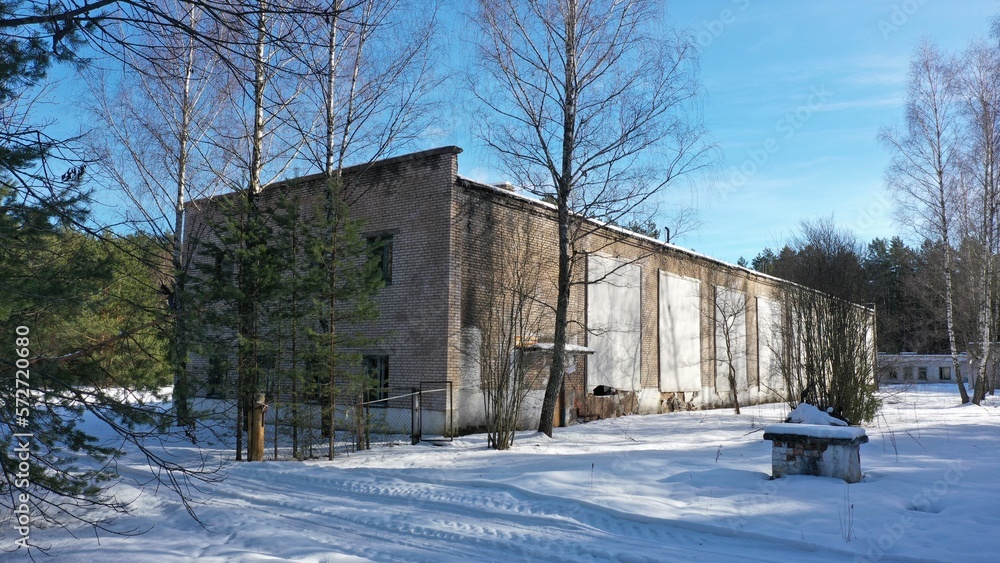 Abandoned building for the storage of military equipment of the former Soviet military base in winter. An old hangar for automotive vehicles under a layer of snow in the middle of a pine forest.