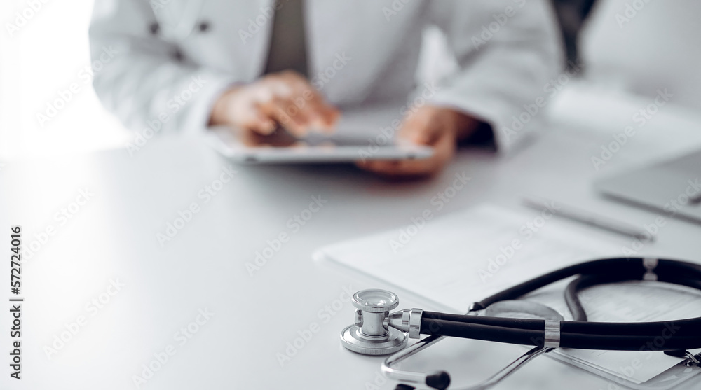 Fototapeta premium Stethoscope lying at doctor's working desk, close up. Physician uses tablet computer at the background. Medicine concept