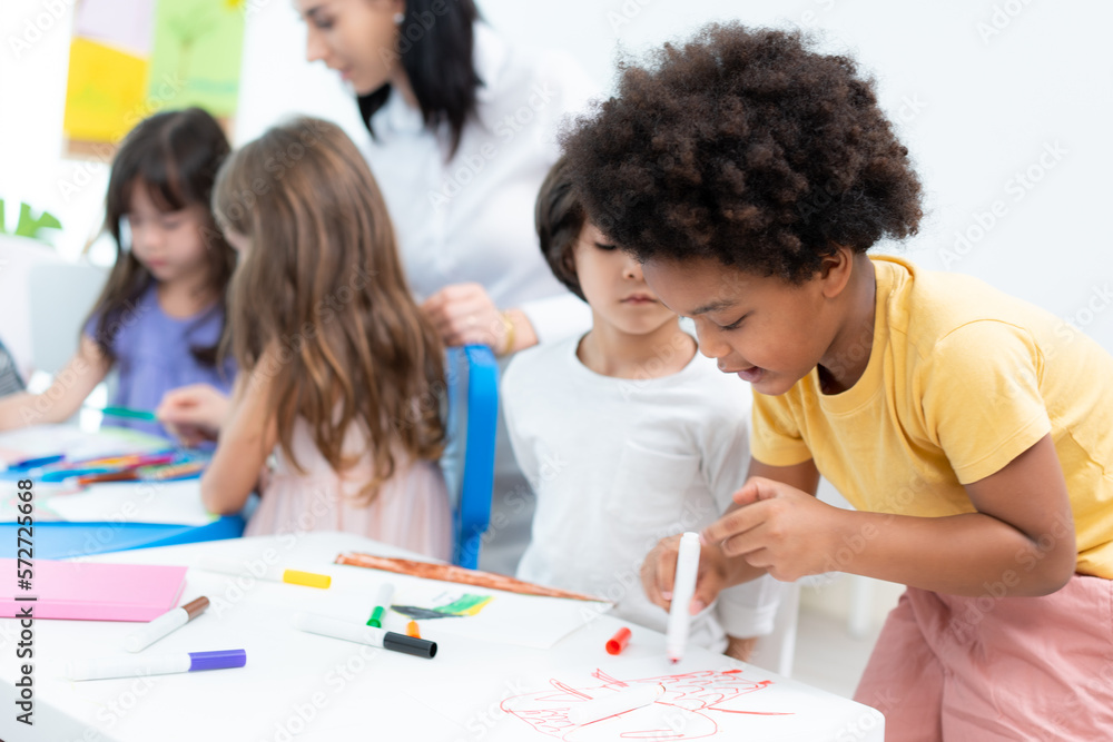 Selective focus of a little African American boy with Afro hair, standing drawing on paper with a red brush pen at a table next to a friend who sitting and watching while drawing at a kindergarten. Selective focus of a little African American boy with Afro hair, standing drawing on paper with a red brush pen at a table next to a friend who sitting and watching while drawing at a kindergarten.