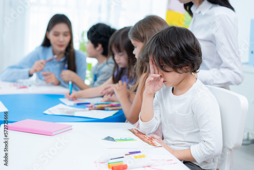 Selective focus of a cute Asian boy sitting at a small table, placed two fingers between his eyebrow head and looking forward, create ideas while drawing with friends in kindergarten classroom