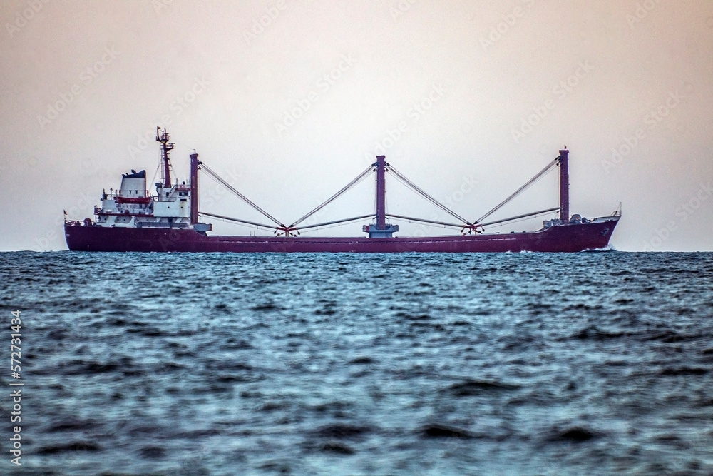 A red ship with antennas and metal poles on board floats on the sea horizon under a cloudy sky