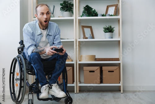 A man in a wheelchair gamer plays games with a joystick in his hands at home, surprise open mouth, copy space, with tattoos on his hands, health concept man with disabilities, real person