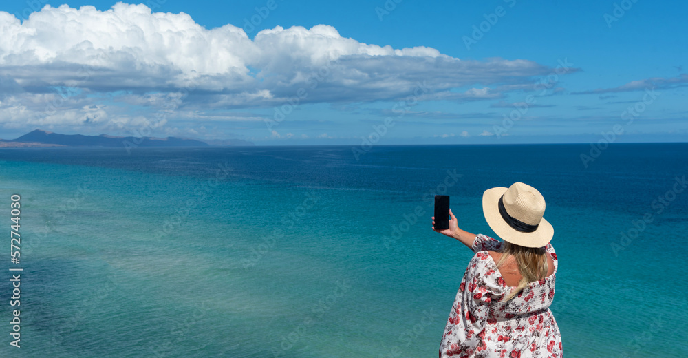 Vista panorámica de una mujer rubia con un sombrero de paja tomando una foto del mar turquesa frente a la costa de la isla turística de Fuerteventura en las Islas Canarias.