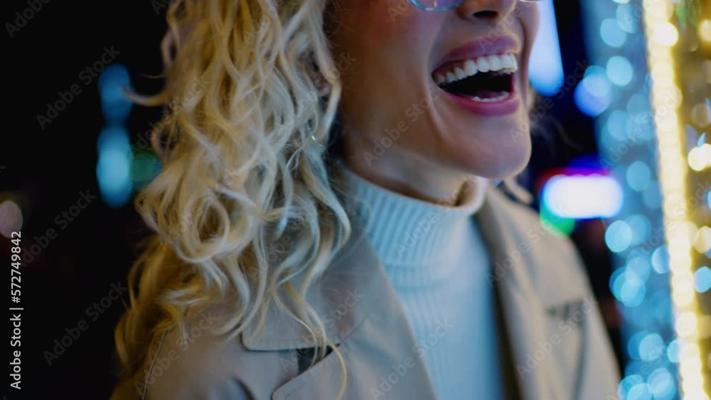 Happy attractive blonde female in stylish glasses stands next to bright glowing lights that are reflected in glass of eye wear. Carefree emotionally laughing young woman enjoying festive feria in city