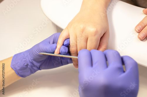 Manicurist shapes, files beautiful woman's nails, holding finger. Caucasian manicure master works in gloves at white work station in spa salon. Soft, safe beauty nail care, treatment. Horizontal plane photo