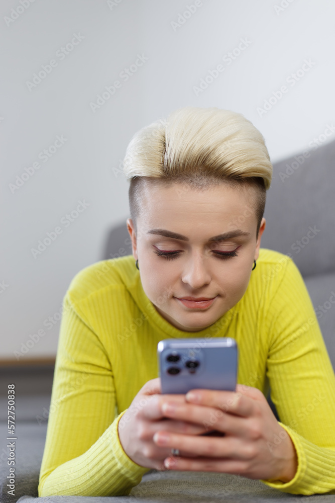 Beautiful young female lying on couch at home and typing a message on smart phone. Stylish short haired tom boy person using modern mobile phone Beautiful young female lying on couch at home and typing a message on smart phone. Stylish short haired tom boy person using modern mobile phone