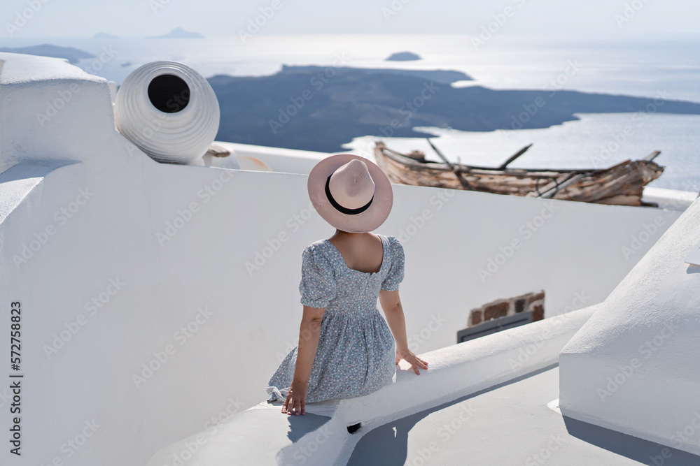Fototapeta premium Tourist woman in blue dress with hat relaxing at sea view in the traditional village Thira. Walking Santorini tour, Greece, during her summer crouise travel. European tourism attraction in Greece.