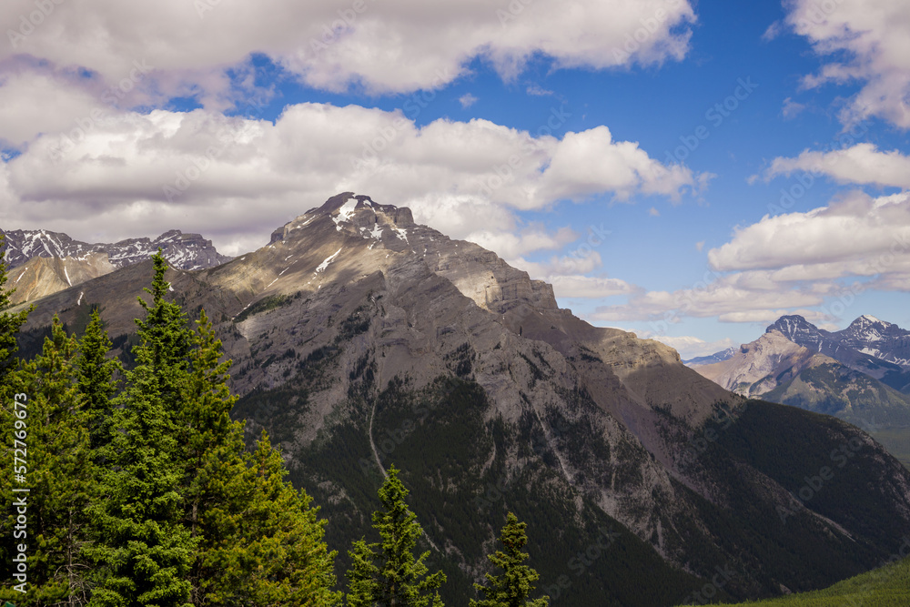 Mountain peaks of the Rocky Mountains in summer. Natural landscape background. 
Coniferous forest, remnants of snow. blue sky and clouds. Tourist season in Banff, Canada