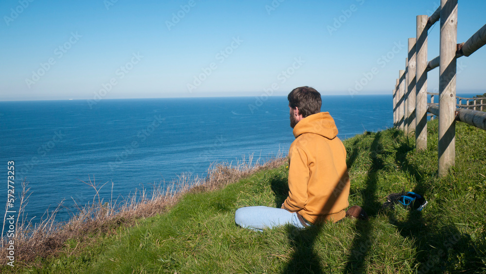Hombre con sudadera sentado en ladera de hierba frente a horizonte marino 