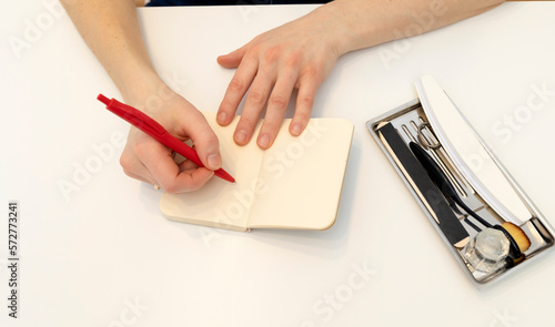 Unrecognizable nail master makes notes in notebook with pen, sitting at workstation,able in nail beauty treatment salon.Professional tools are on white table.Manicurist's routine.Top view Horizontal photo