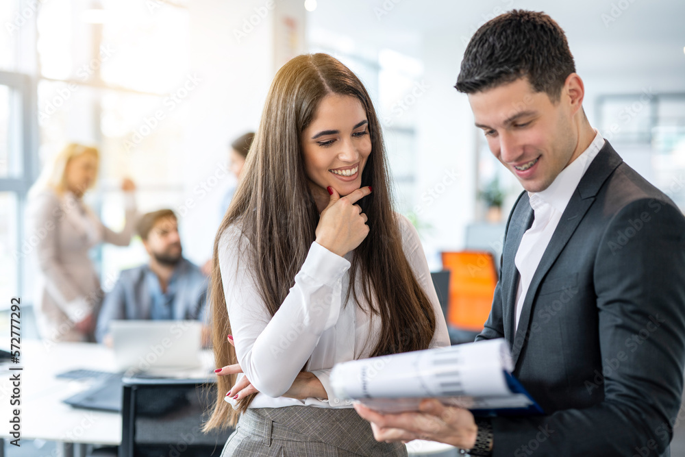 Attractive young woman in formal wear checking financial reports and paperwork with handsome good looking man in suit while standing in office. Attractive young woman in formal wear checking financial reports and paperwork with handsome good looking man in suit while standing in office.
