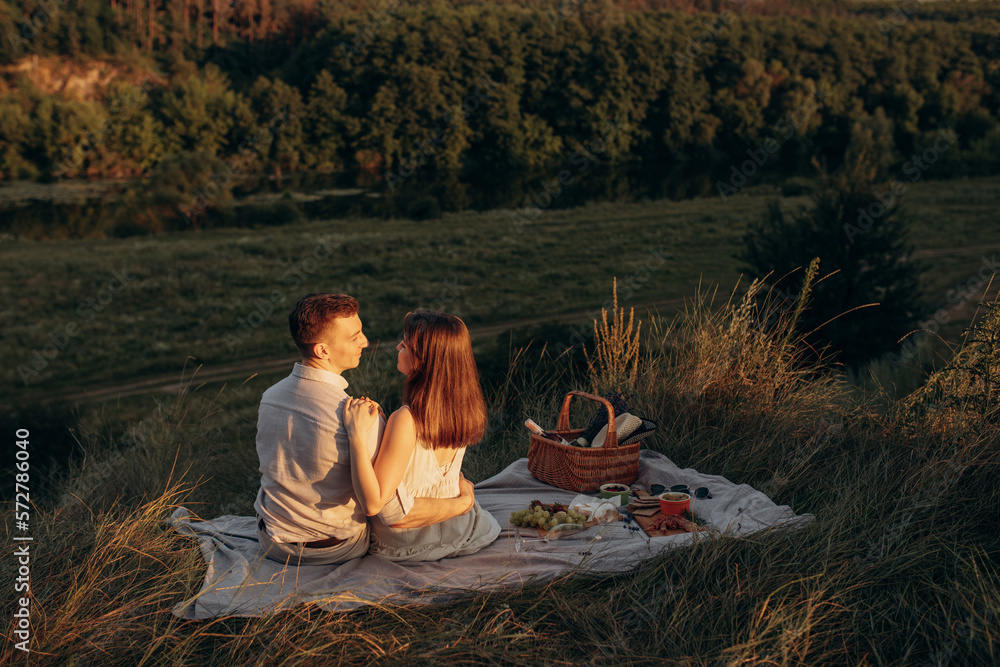 A couple in love, husband and wife, are sitting on a picnic on the mountain and admiring nature. Lovestory at sunset in summer. Warm family shooting in nature