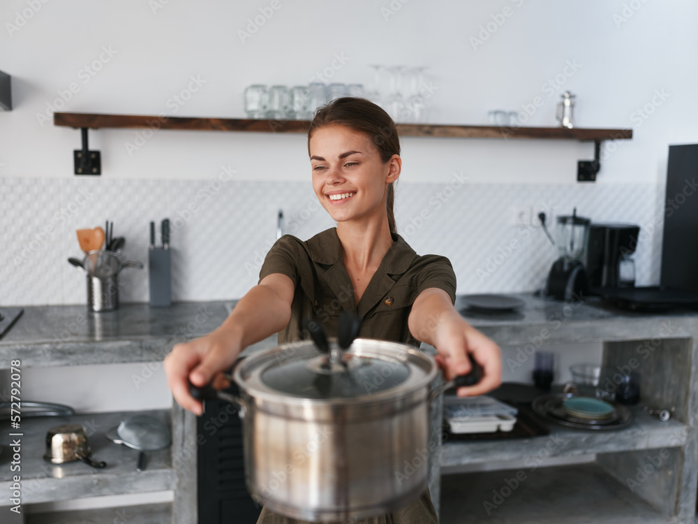A woman in stylish kitchen prepares food and holds a cooking pot in her hands for cooking dinner, camera and smiles with teeth, looking into the camera. Home atmosphere, lifestyle, comfort A woman in stylish kitchen prepares food and holds a cooking pot in her hands for cooking dinner, camera and smiles with teeth, looking into the camera. Home atmosphere, lifestyle, comfort