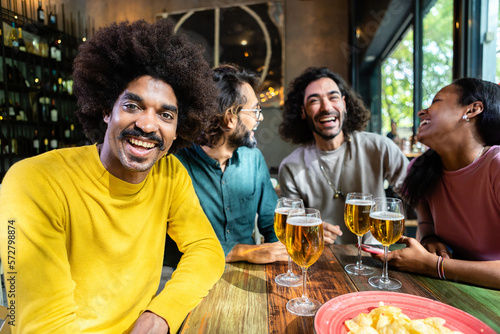 Group of happy young adult friends toasting beer at happy hours after work. Colleagues people having fun together at brewery bar. Smiling african american man smiling at camera at pub.