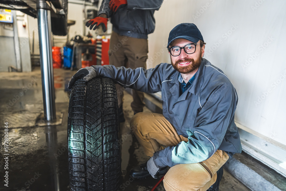 Shot of a mechanic squatting down, smiling and holding one hand on a car tyre. High quality photo