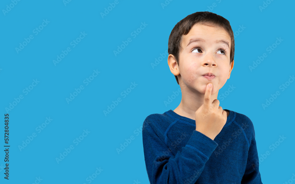 Pensive kid looks away at copy space thinking isolated on a blue background, funny kid lips hold finger near mouth conceiving some kind of joke, conceptual image Pensive kid looks away at copy space thinking isolated on a blue background, funny kid lips hold finger near mouth conceiving some kind of joke, conceptual image