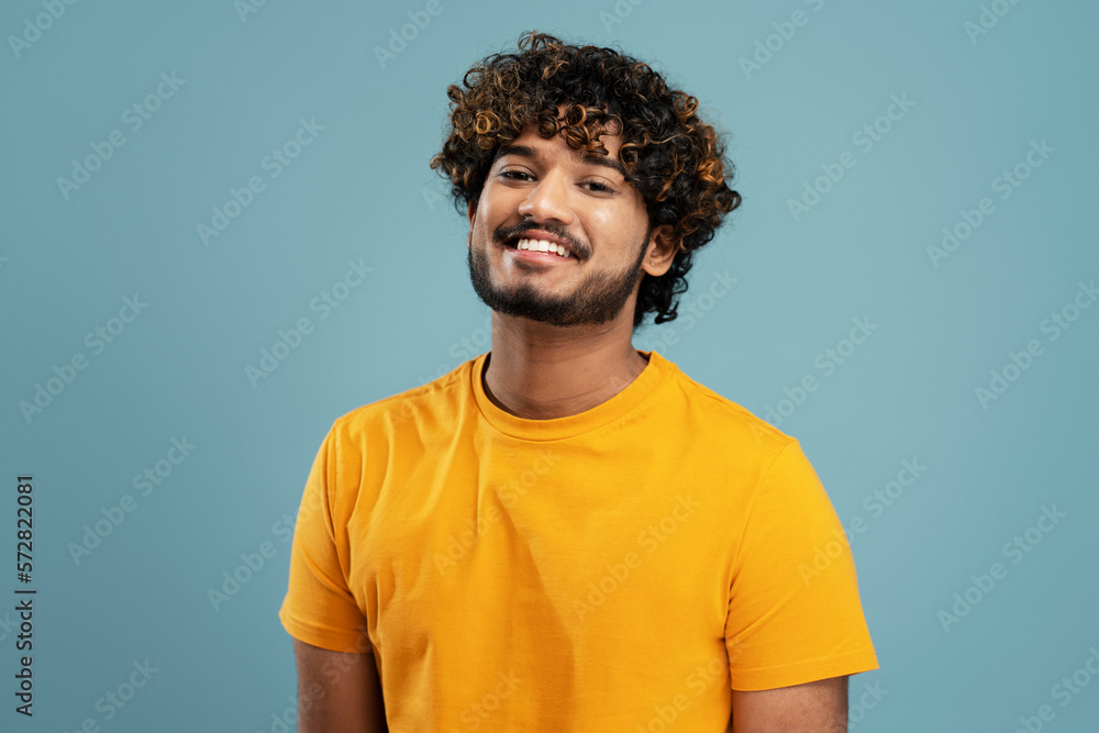 Confident curly haired bearded Indian man, wearing a bright yellow t-shirt, smiling a beautiful toothy smile looking at camera, isolated over blue background Confident curly haired bearded Indian man, wearing a bright yellow t-shirt, smiling a beautiful toothy smile looking at camera, isolated over blue background