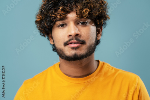 Cropped portrait of a hipster Indian man in yellow t-shirt, confidently looking at camera, over isolated blue background © Maria Vitkovska