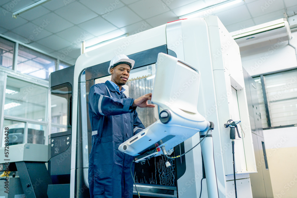 Lower and front view of African American worker do some works with keyboard and monitor of controlling part of factory robot machine with safety protection cloth and hat and look happiness workplace.