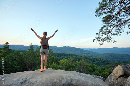 Sportive woman standing alone on hillside trail with raised up arms. Female hiker enjoying view of evening nature from rocky cliff on wilderness path. Active lifestyle concept