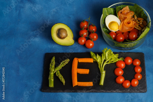 glass bowl with cherry tomato, avocado, egg, salmon and nuts with a sign with the word keto