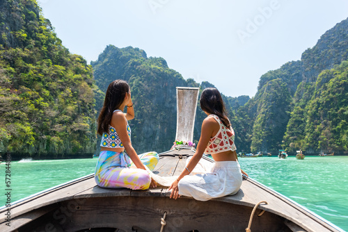 Asian woman friends sitting on ship bow and looking beautiful island beach lagoon during travel on boat in summer sunny day. Attractive girl enjoy and fun outdoor lifestyle travel on holiday vacation