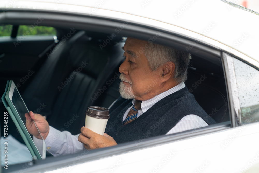 Confident senior business man in suit sitting on car backseat with working on digital tablet and drinking hot coffee during go to office. Elderly businessman executive working on the road.