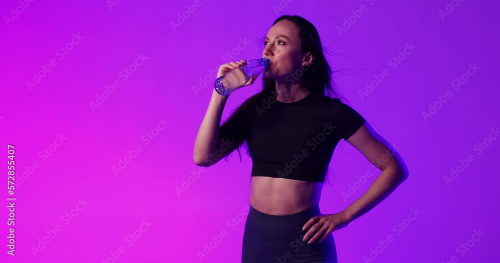 Portrait of an athletic young woman in sports clothes drinking a water from bottle, preparing for training in a studio. Beautiful muted purple lighting. Active lifestyle concept. Portrait of an athletic young woman in sports clothes drinking a water from bottle, preparing for training in a studio. Beautiful muted purple lighting. Active lifestyle concept.