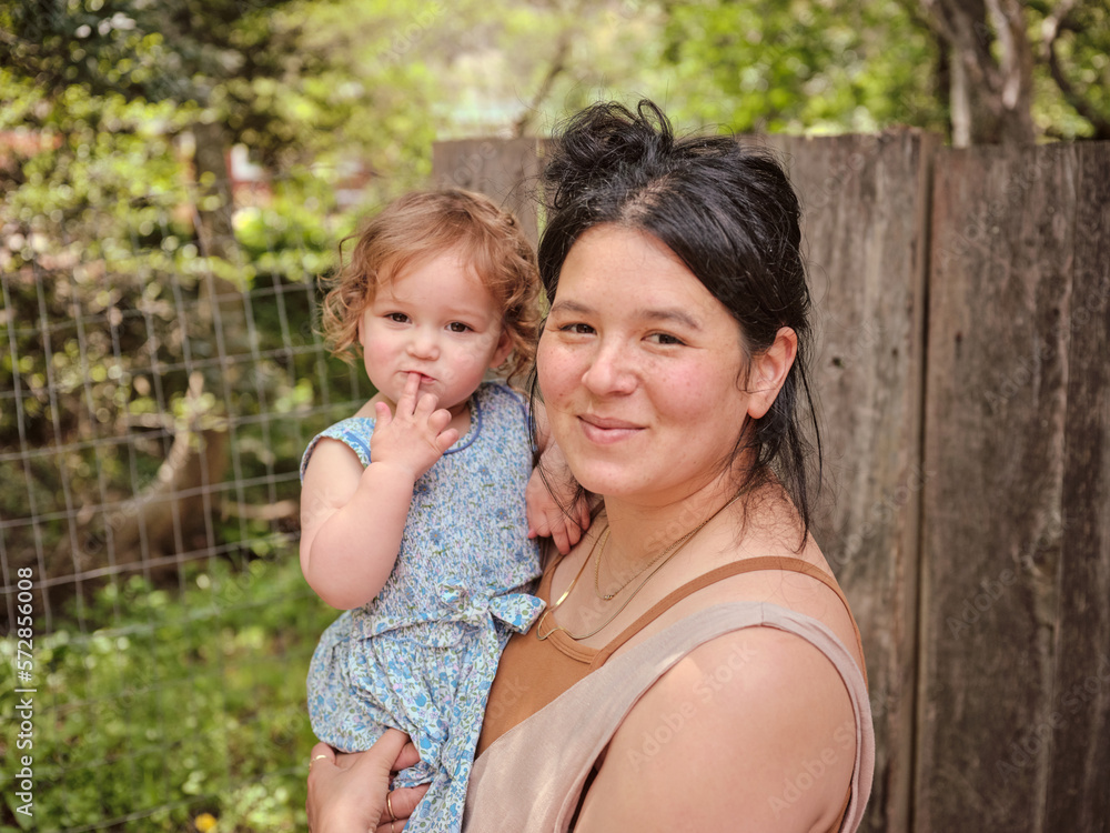 Mom holding daughter and looking at camera in natural backyard Mom holding daughter and looking at camera in natural backyard