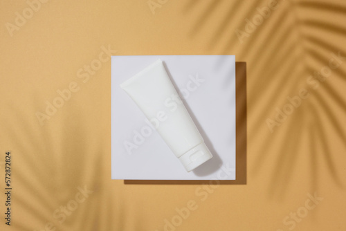 Sunscreen products concept. Top view photo of cosmetic cream tube on square white podium and tropical leaves shadow on sandy background. Flat lay cosmetics mockup idea.