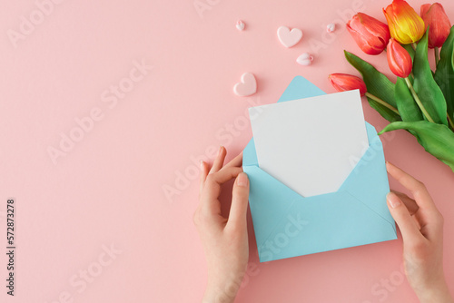 Woman day concept. First person top view photo of female hands holding open envelope with letter and tulips flowers hearts on pastel pink background with blank space. Holiday card idea.