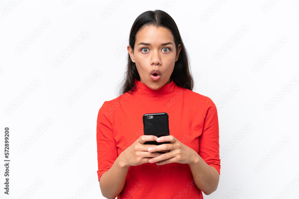 Young Colombian woman isolated on white background looking at the camera while using the mobile with surprised expression Young Colombian woman isolated on white background looking at the camera while using the mobile with surprised expression