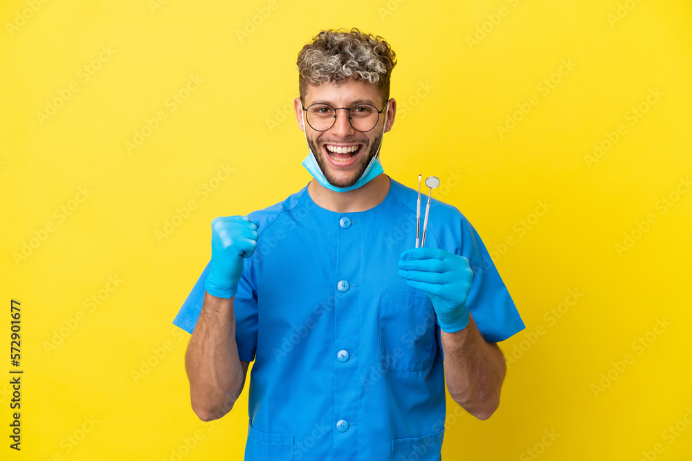 Dentist caucasian man holding tools isolated on yellow background celebrating a victory in winner position Dentist caucasian man holding tools isolated on yellow background celebrating a victory in winner position