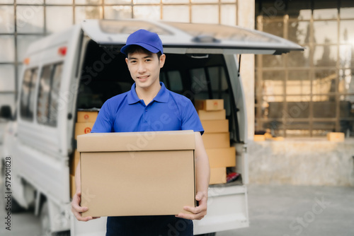 Asian delivery man portrait of young man holding cardboard box while unloading moving van outdoors. © NINENII