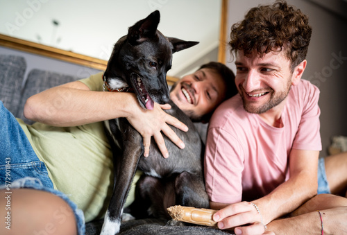 Homosexual couple on the sofa playing whit dog at cozy home. Concept of homosexual relations and lifestyle at home. Idea of  gay family concept