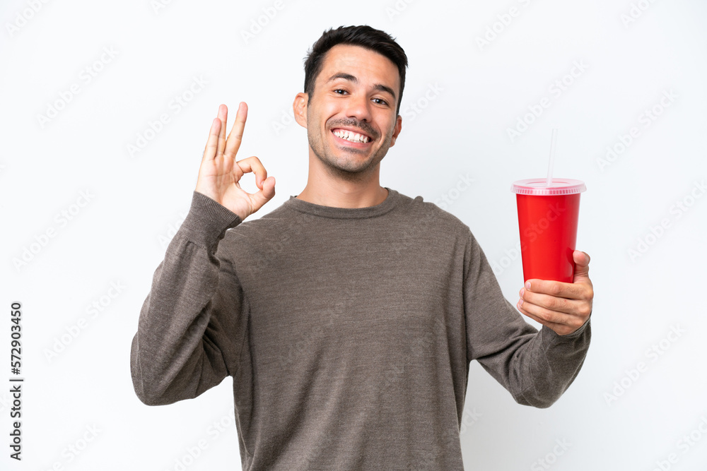 Young handsome man holding soda over isolated white background showing ok sign with fingers Young handsome man holding soda over isolated white background showing ok sign with fingers