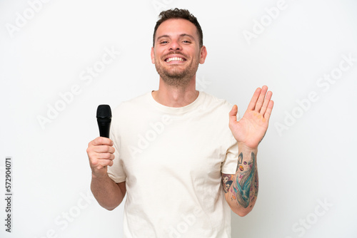 Young caucasian singer man picking up a microphone isolated on white background saluting with hand with happy expression