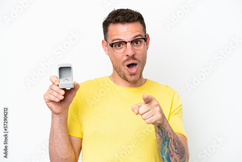 Young caucasian man holding a engagement ring isolated on white background surprised and pointing front