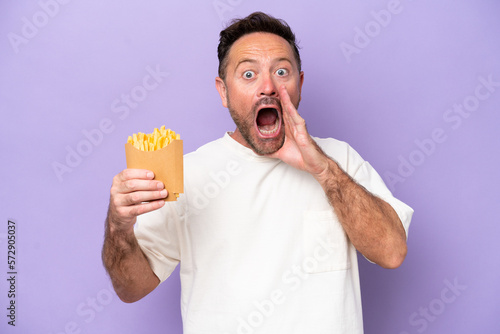 Middle age caucasian man holding fried chips isolated on purple bakcground with surprise and shocked facial expression © luismolinero