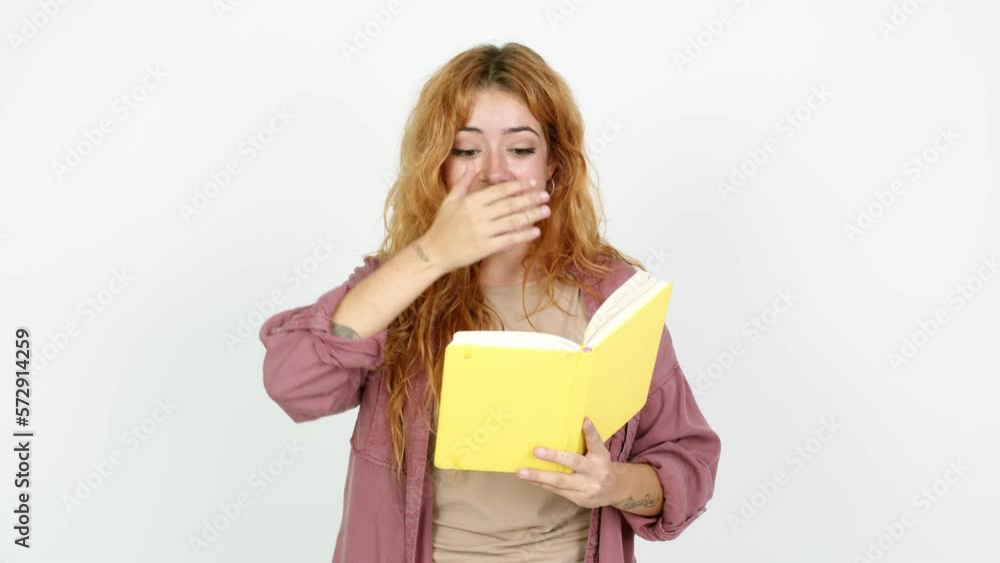 Young redhead woman surprised and holding a book over isolated background Young redhead woman surprised and holding a book over isolated background