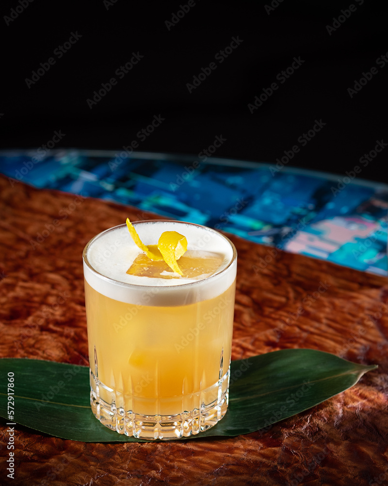 Cocktail at the bar on brown leather background. Whisky amaretto sour with lemon zest garnishment. Modern, stylish alcoholic drink at restaurant. Beautiful beverage mix made with bourbon. Vertical