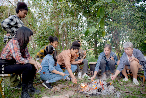Happy friends group doing recreation with man playing guitar while friends and children grill marshmallow at camping together in holiday, tourist and picnic, party bonfire in vacation.