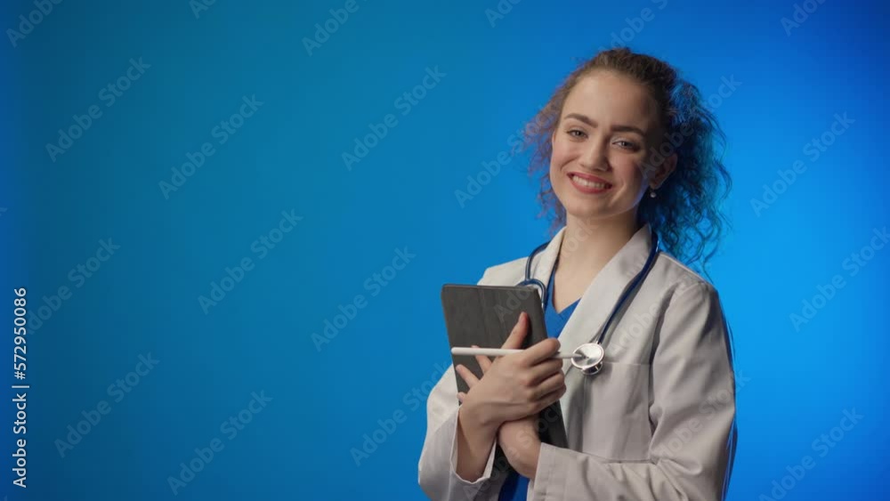Studio videoshot of a young female doctor looking at camera against a blue background. Studio videoshot of a young female doctor looking at camera against a blue background.