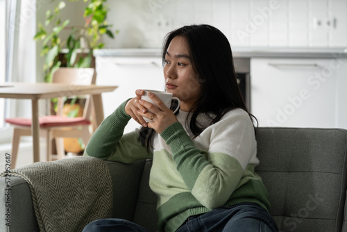 Relaxed pensive Asian woman in casual clothes sits on sofa with cup of hot coffee in hands. Young introverted japanese brunette lady looking into distance enjoying being alone in own apartment