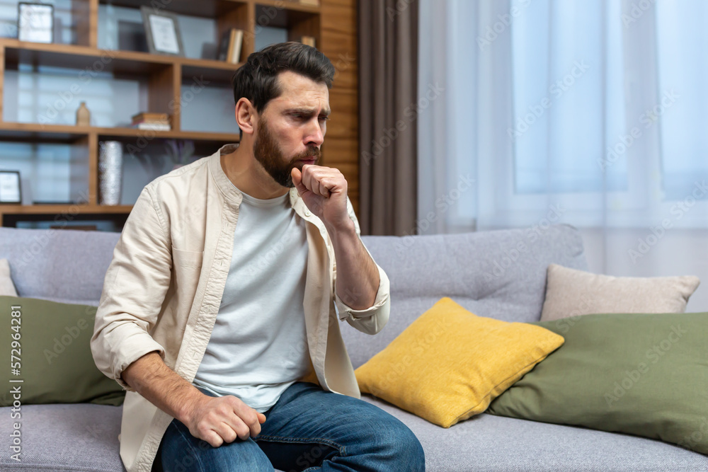 Sick mature man alone at home sitting on sofa coughing holding hands to chest in living room. Sick mature man alone at home sitting on sofa coughing holding hands to chest in living room.