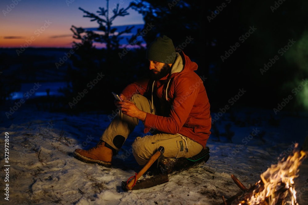 A hiker checks his phone while warming himself by a campfire at night in the mountains. A hiker checks his phone while warming himself by a campfire at night in the mountains.