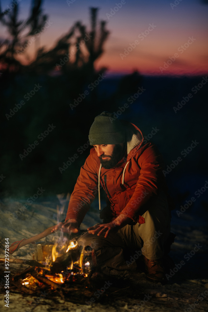 A man sits near a campfire, warming himself by the fire in a winter forest at sunset. A man sits near a campfire, warming himself by the fire in a winter forest at sunset.